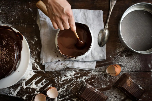 Top view of a man’s hand mixing chocolate in a bowl with a chocolate cake on the left and a spoon, sifter, chocolate bars, egg shells, and sifted flour around.