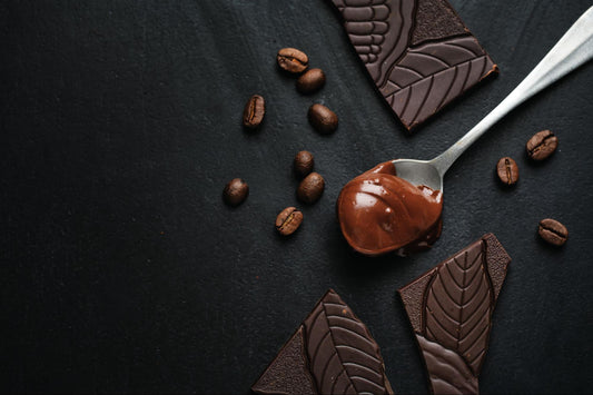 A spoon of melted chocolate next to cocoa beans and broken chocolate bars with a leafy design showcasing chocolate's rich textures and natural origins.