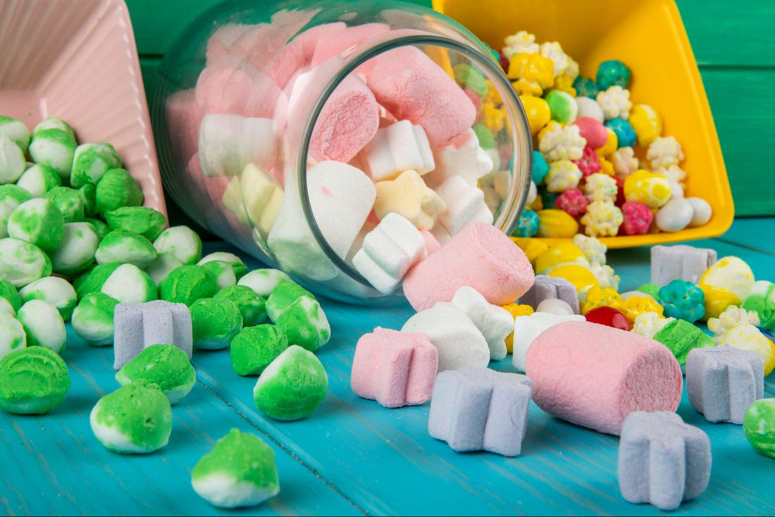 A side view of various colorful candies and marshmallows scattered from bowls and a glass jar on a green wooden background.