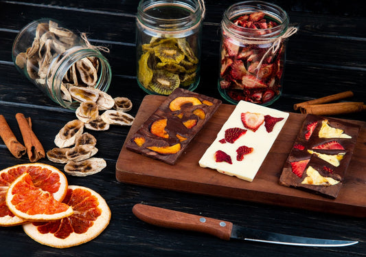 Side view of dark and white chocolate bars with embedded dried fruits displayed on a wooden cutting board. Bottles filled with assorted dried fruits surround the chocolate, and a knife is placed in the foreground.